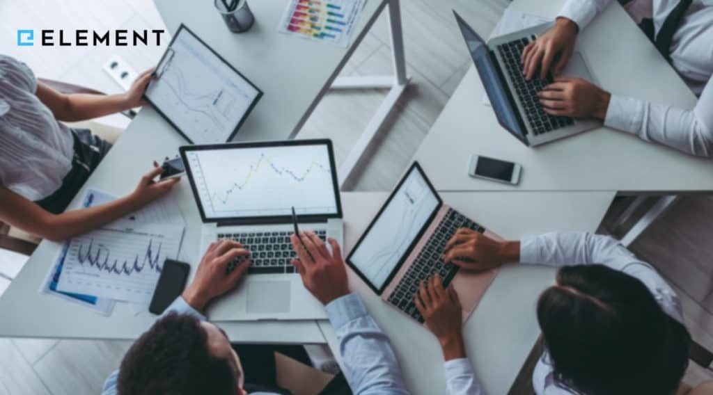 Ilustracja do artykułu o historii systemu Element. Przedstawia pracowników siedzących w kółko z laptopami na stole. Zdjęcie zrobione jest z perspektywy ptaka. W lewym górnym rogu znajduje się logo Element.