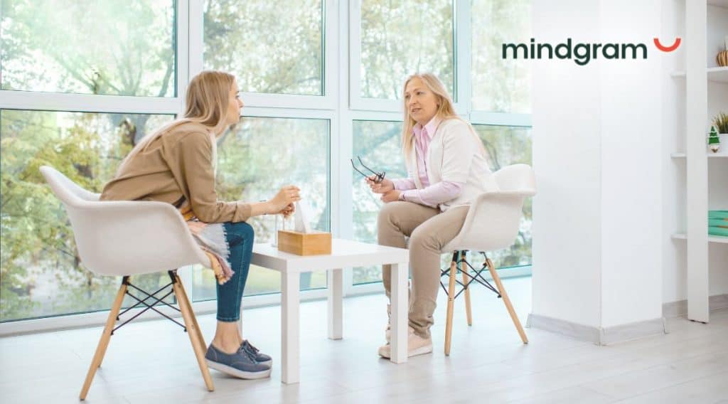 Zdjęcie przedstawia 2 kobiety, jest to rozmowa psycholożki z pacjentką. W górny prawym rogu znajduje się logo: Mindgram.