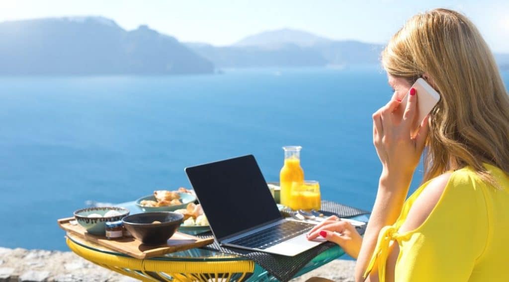 Zdjęcie jest ilustracją do artykułu: Nielimitowane urlopy. Strategia PR-owa czy realna poprawa dobrostanu pracowniczego?. Przedstawia młdoą kobietę siedząca przy stoliku na którym satoi laptop, przekąski i sok pomarańczowy. Kobieta rozmawia przez telefon. Przed nią rozciąga się widok na morze i wyspy.
