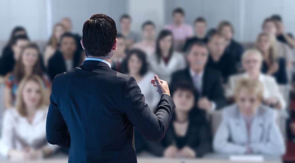 Zdjęcie jest ilustracją do artykułu: Jaki mindset powinien mieć założyciel startupu rekrutacyjnego, aby odniósł sukces?. Przedstawia mężczyznę w garniturze, stojącego tyłem do czytelnika i przemawiającego przed widownią złożoną z ludzi w różnym wieku i różnej płci.