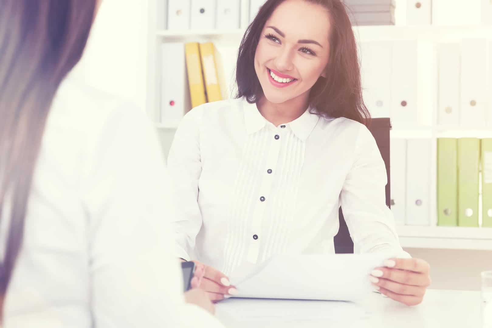 Zdjęcie jest ilustracją do artykułu: Spotkanie Kobiet HR w Rzeszowie. Przedstawia uśmiechniętą młodą kobietę z działu HR, siedzącą przy biurku i rozmawiającą z kandydatką.