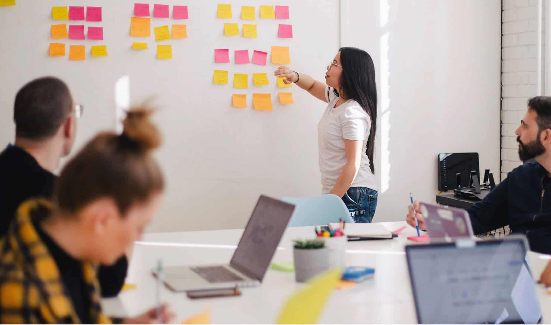 Zdjęcie jest ilustracją artykułu "Jak Agile może pomóc HR?". Przedstawia spotkanie grupy roboczej pracowników. Jedna kobieta stoi przy tablicy pełnej kolorowych post it. Pozostałe osoby z zespołu siedza przy stole konferencyjnym