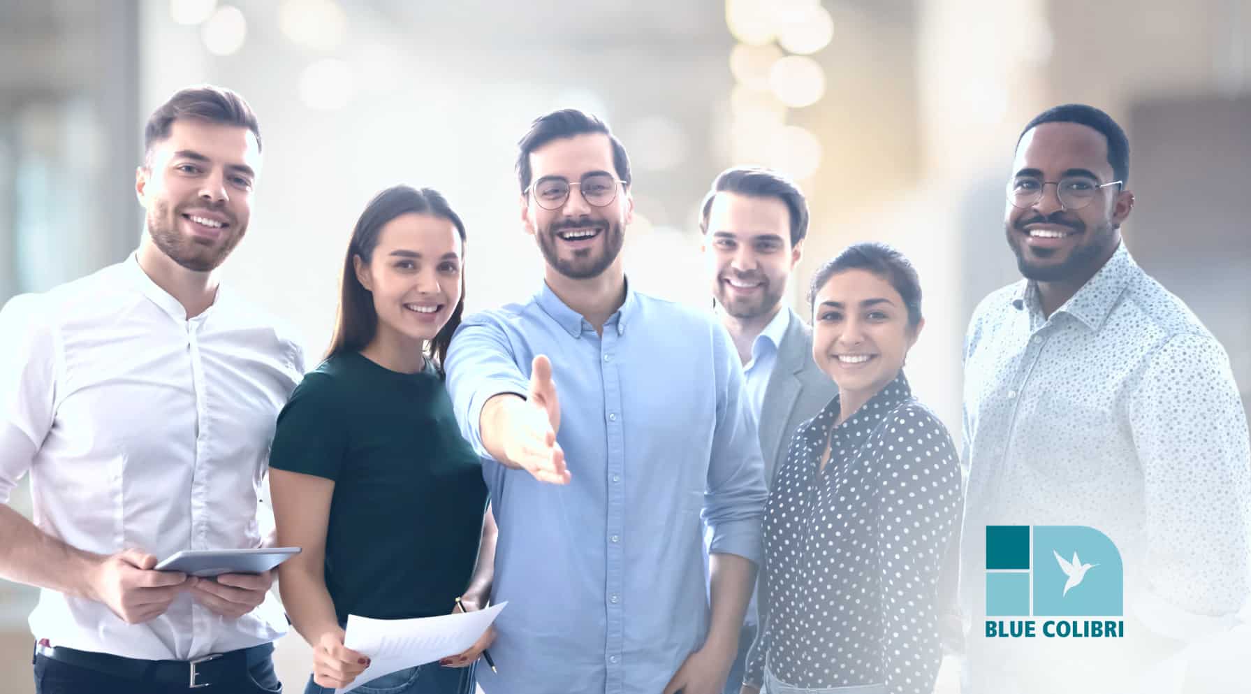 Zdjęcie jest ilustracją do artykułu : Czego potrzebuje pracownik właśnie przyjęty do pracy? O sprawnym preboardingu i onboardingu, Przedstawia grupę młodych uśmiechniętych pracowników, z których jeden wyciąga rękę w kierunku patrzącego w geście powitania. W dolnym prawym rogu logo Blue Colibri.