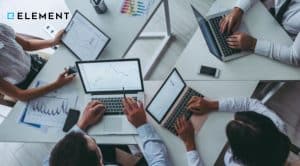 Ilustracja do artykułu o historii systemu Element. Przedstawia pracowników siedzących w kółko z laptopami na stole. Zdjęcie zrobione jest z perspektywy ptaka. W lewym górnym rogu znajduje się logo Element.