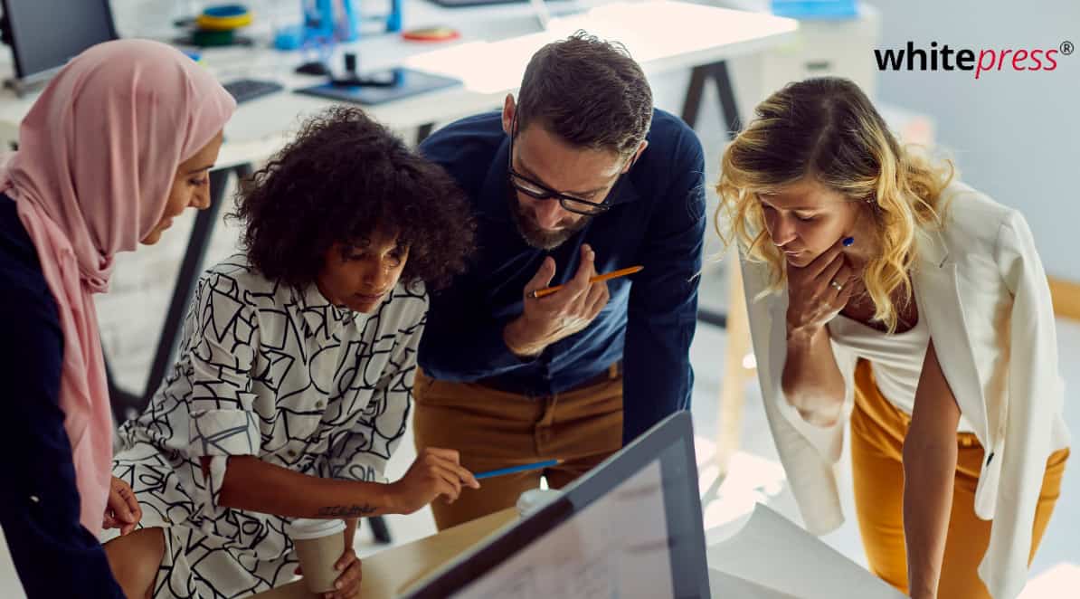 Zdjęcie jest ilustracją do artykułu: Globalny raport 2022 o diversity. Przedstawia grupę pracowników z różnych kultur. Cztery osoby pochylone są nad stołem i dyskutują nad czymś. W prawym górnym rogu logo WhitePress.
