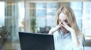 Zdjęcie jest ilustracją do artykułu: Atmosfera uwikłania – subtelna manipulacja. Przedstawia młodą kobietę przed laptopem, która ma opuszczoną głowę, zamknięte oczy a koniuszkami palców podtrzymuje czoło. Jej postawa sugeruje przeżywanie silnego stresu.