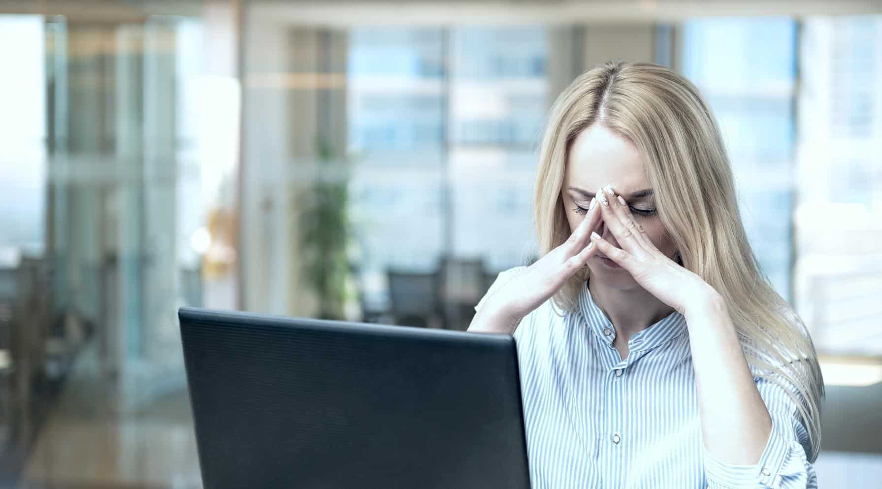 Zdjęcie jest ilustracją do artykułu: Atmosfera uwikłania – subtelna manipulacja. Przedstawia młodą kobietę przed laptopem, która ma opuszczoną głowę, zamknięte oczy a koniuszkami palców podtrzymuje czoło. Jej postawa sugeruje przeżywanie silnego stresu.