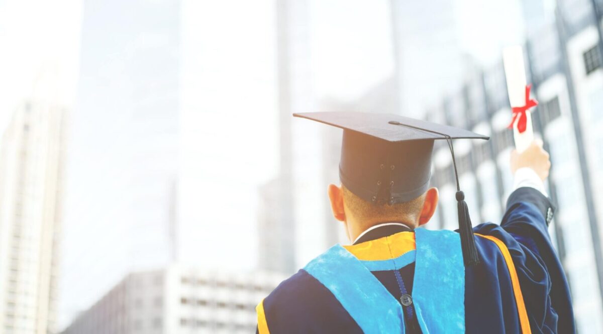 Ilustracja do artykułu: Rankingi Uczelni Wyższych. Przedstawia absolwenta uczelni w uroczystym stroju na tle biurowców.
